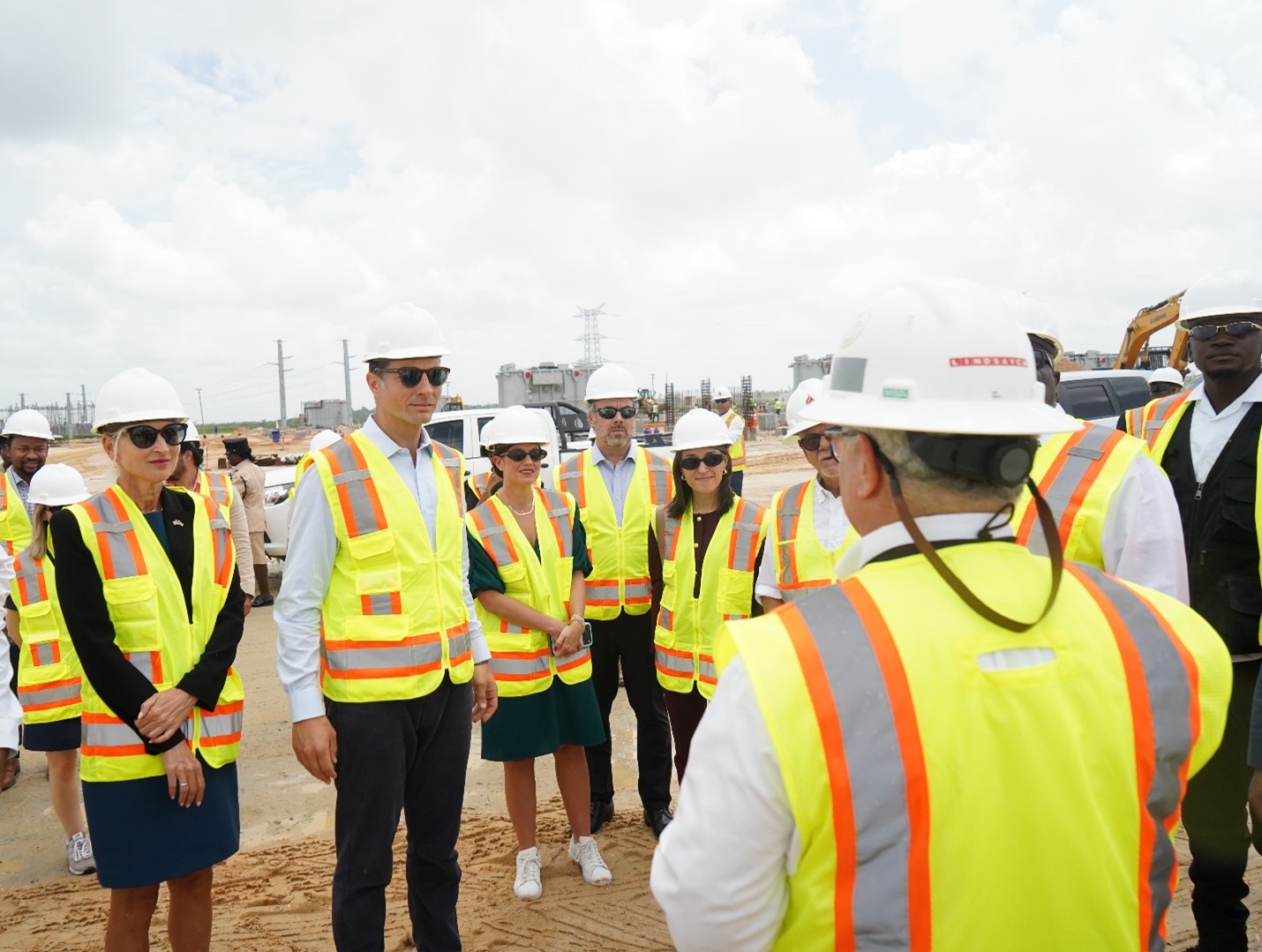 A group of people wearing hard hats and high-visibility safety vests stand on a construction site, listening to a speaker in the foreground who appears to be giving instructions or a briefing.