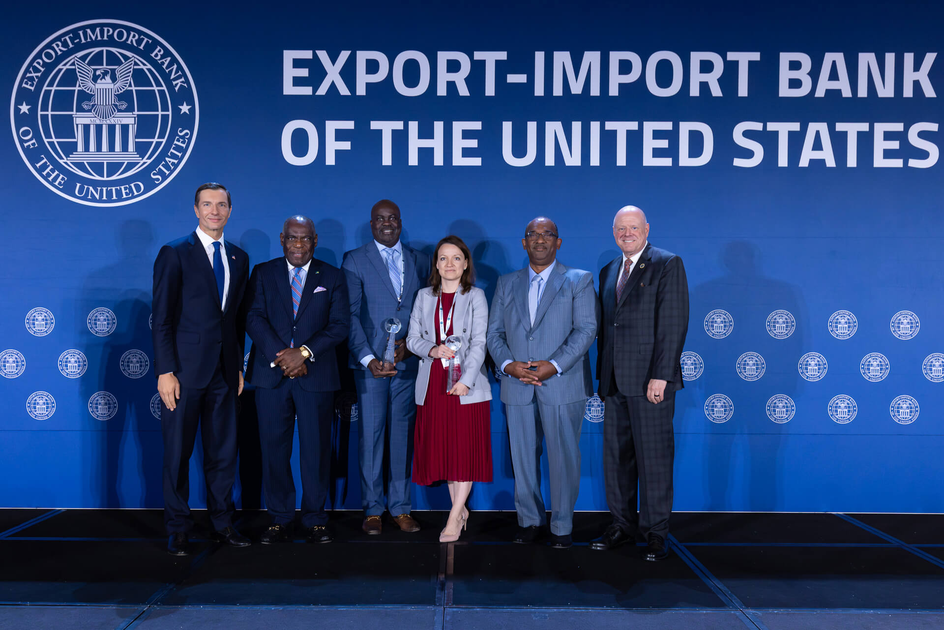 A group of people standing in front of a backdrop containing the EXIM Seal and says, "EXPORT-IMPORT BANK OF THE UNIITED STATES