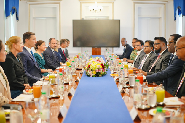 Two groups of formally dressed professionals sit across from each other at a long conference table with drinks and a floral centerpiece, engaged in a formal meeting or discussion in a well-lit room.